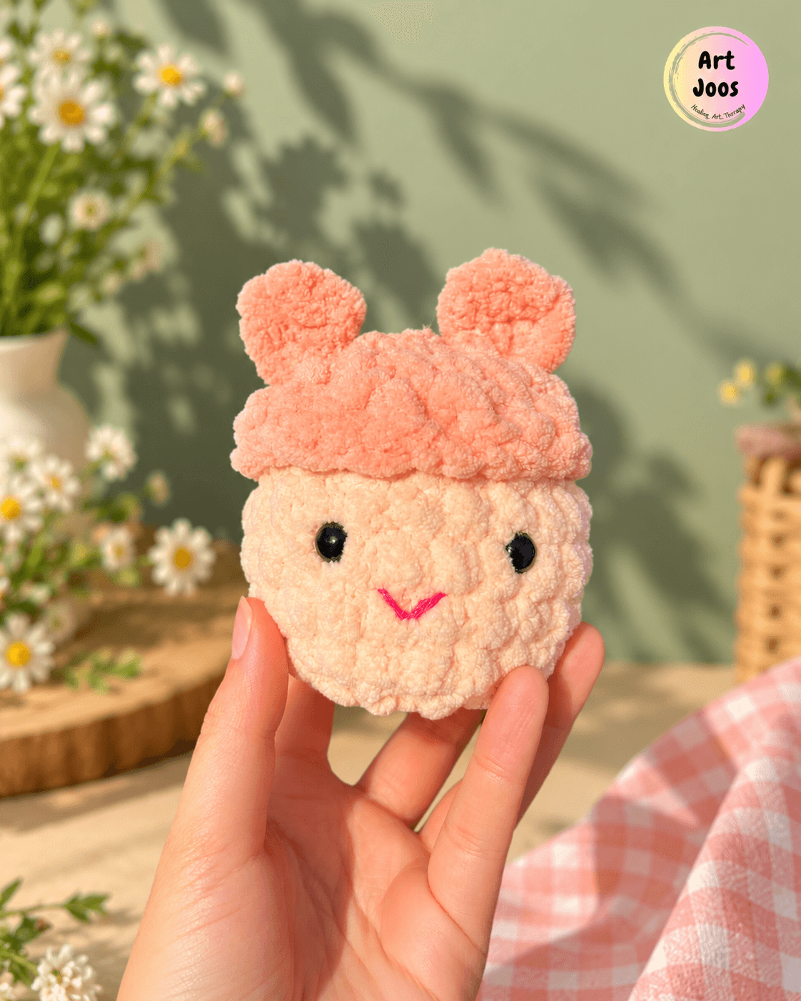 Handmade crochet plushie with a cream-colored round face, peach-pink cap and rounded ears, glossy black eyes, and a tiny pink embroidered smile.