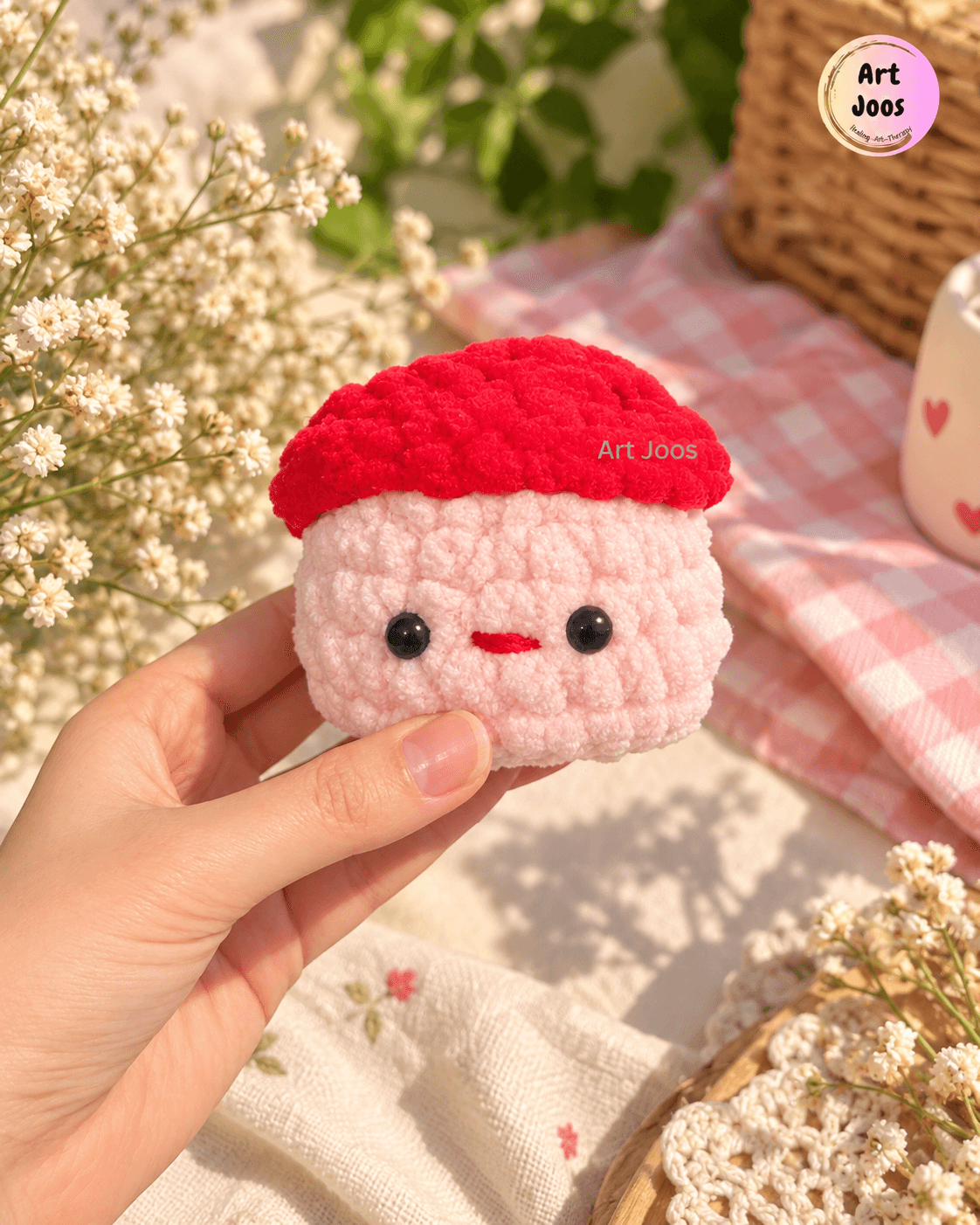 Handmade blush-pink crochet plushie with a bright red textured top, glossy black eyes, and a tiny embroidered smile, held in a hand.