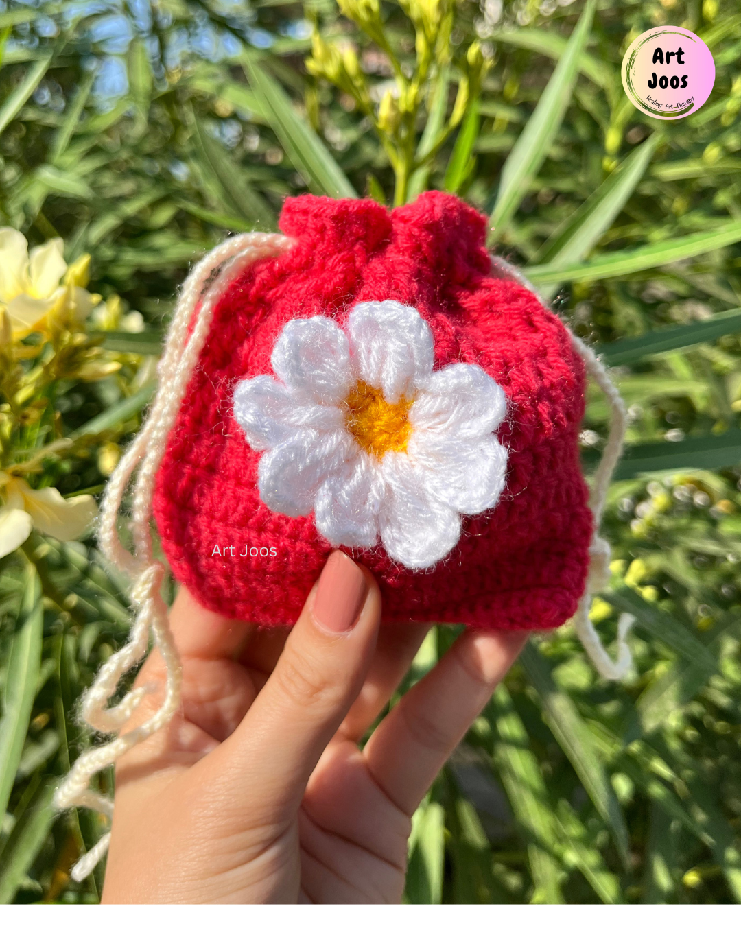 Bright red handmade crochet pouch with a white daisy featuring a yellow center and a cream drawstring closure, styled on a picnic-inspired surface.