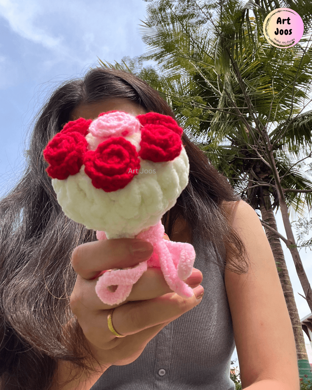 Handmade crochet flower bouquet with four deep red roses, a small pink flower, a fluffy white base, and dangling pink yarn stems, held in a hand.