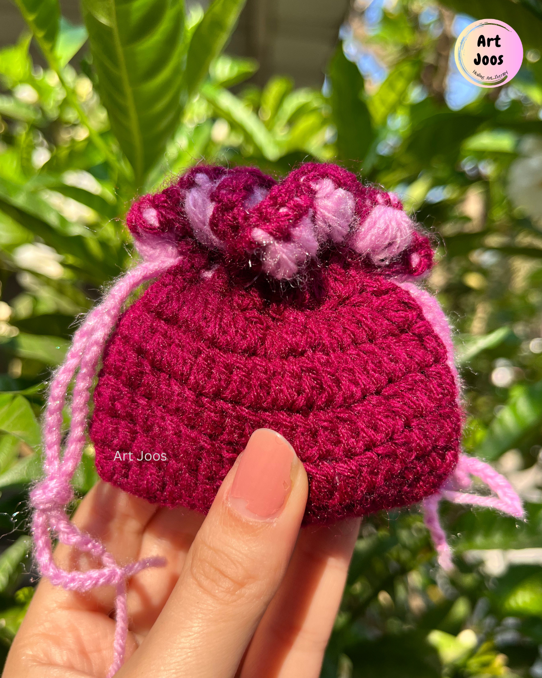 Berry-pink handmade crochet drawstring pouch with a pastel pink scalloped top trim and matching drawstrings, held in a hand.